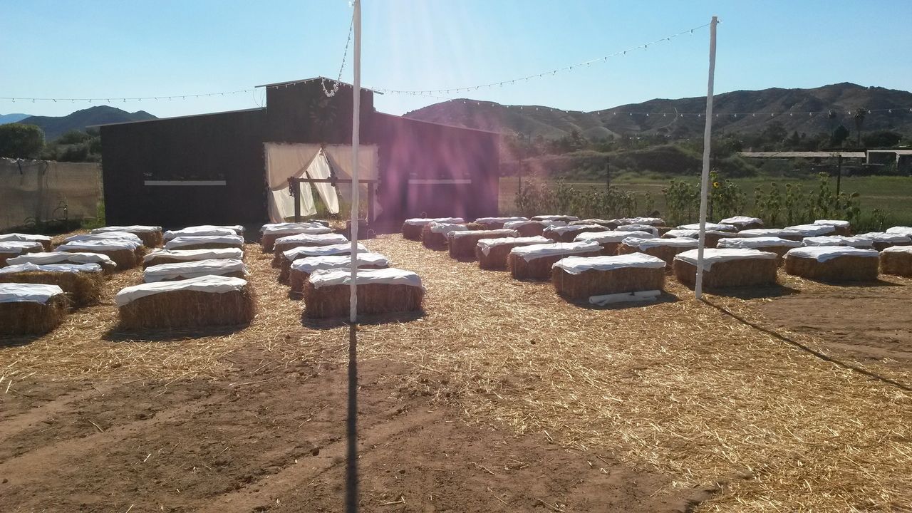Rows of hay bales covered with white cloths are arranged as seating in front of a barn on a sunny day, with string lights overhead and mountains in the background.