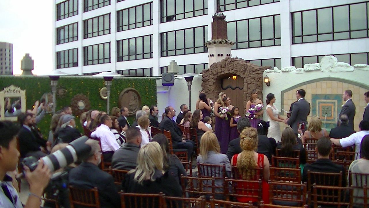 A wedding ceremony takes place outdoors in front of a building, with guests seated and bridesmaids in purple dresses standing beside the couple at the altar.