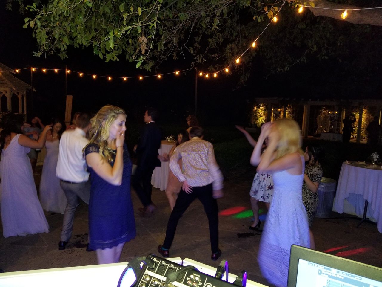 People dancing outdoors at night under string lights, with DJ equipment visible in the foreground and tables in the background.