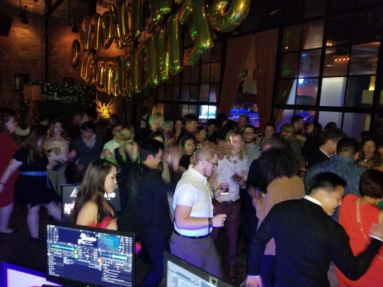 A large group of people are dancing and socializing in a dimly lit venue with gold balloons and DJ equipment visible in the foreground.