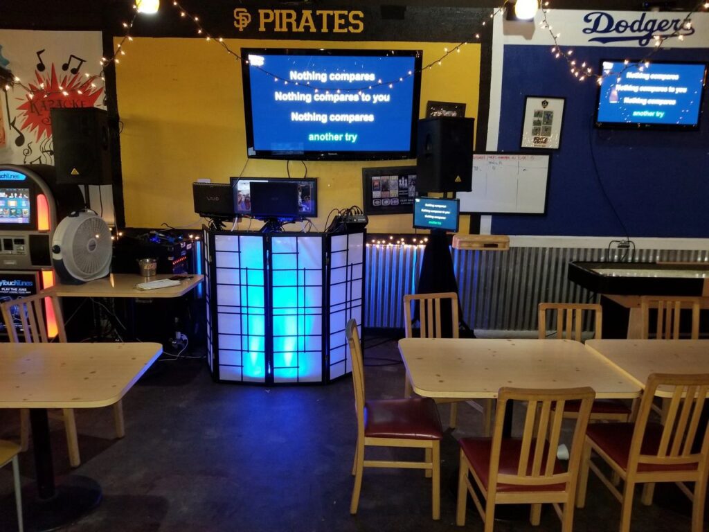 A karaoke setup in a casual dining area with lyrics on TV screens, wooden tables and chairs, and sports team signs on the walls.