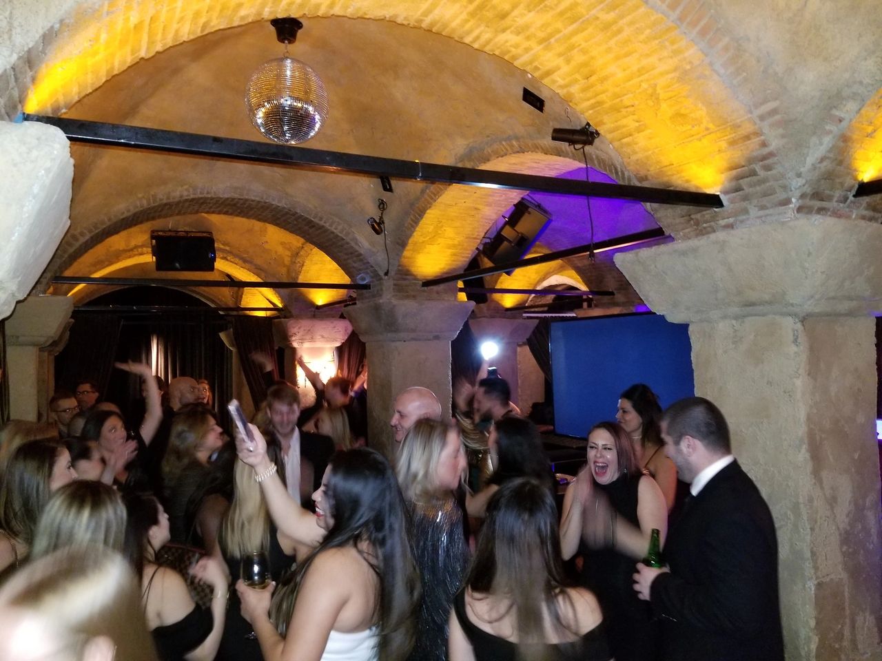 Entrepreneur's Karaoke Dj Party. Guests dressed in formal attire are dancing and socializing at a lively party in a stone-arched room with yellow lighting and a disco ball.