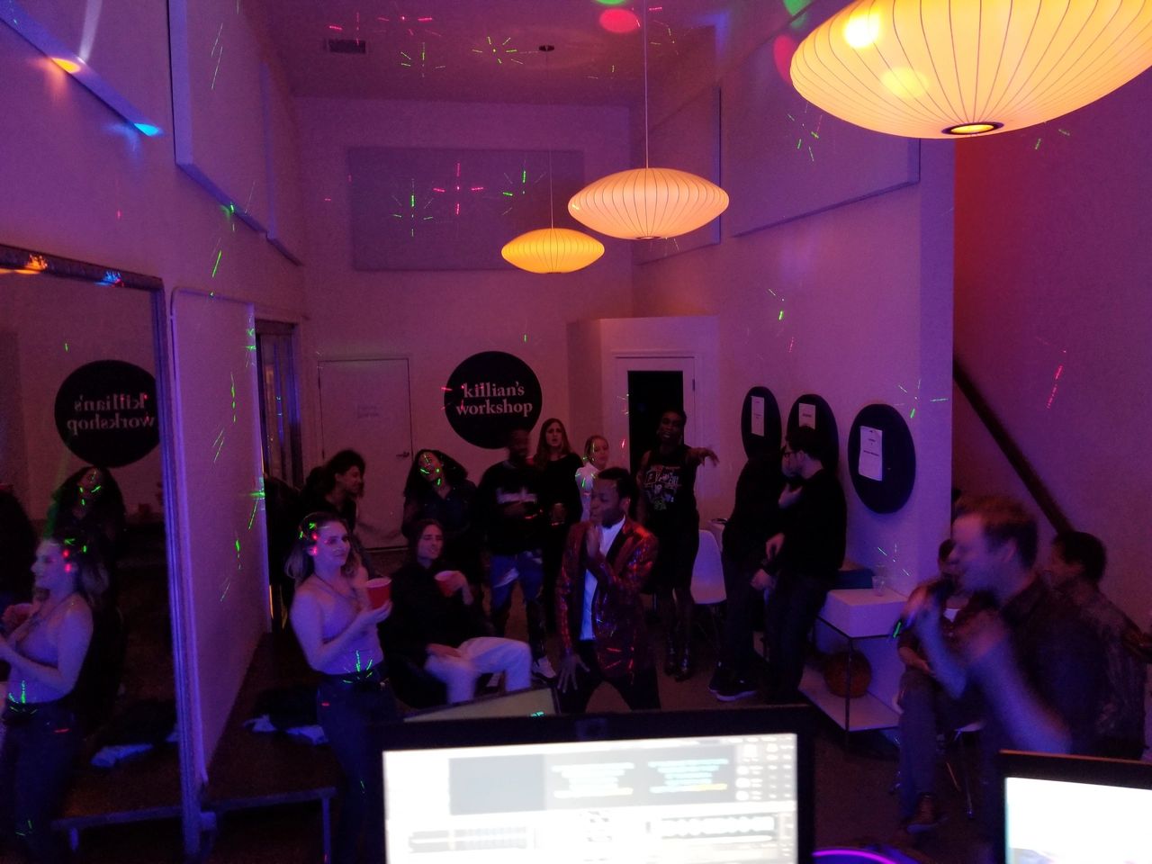 A group of people socialize and dance in a dimly lit room with colorful lights and hanging lamps; computer monitors are visible in the foreground.