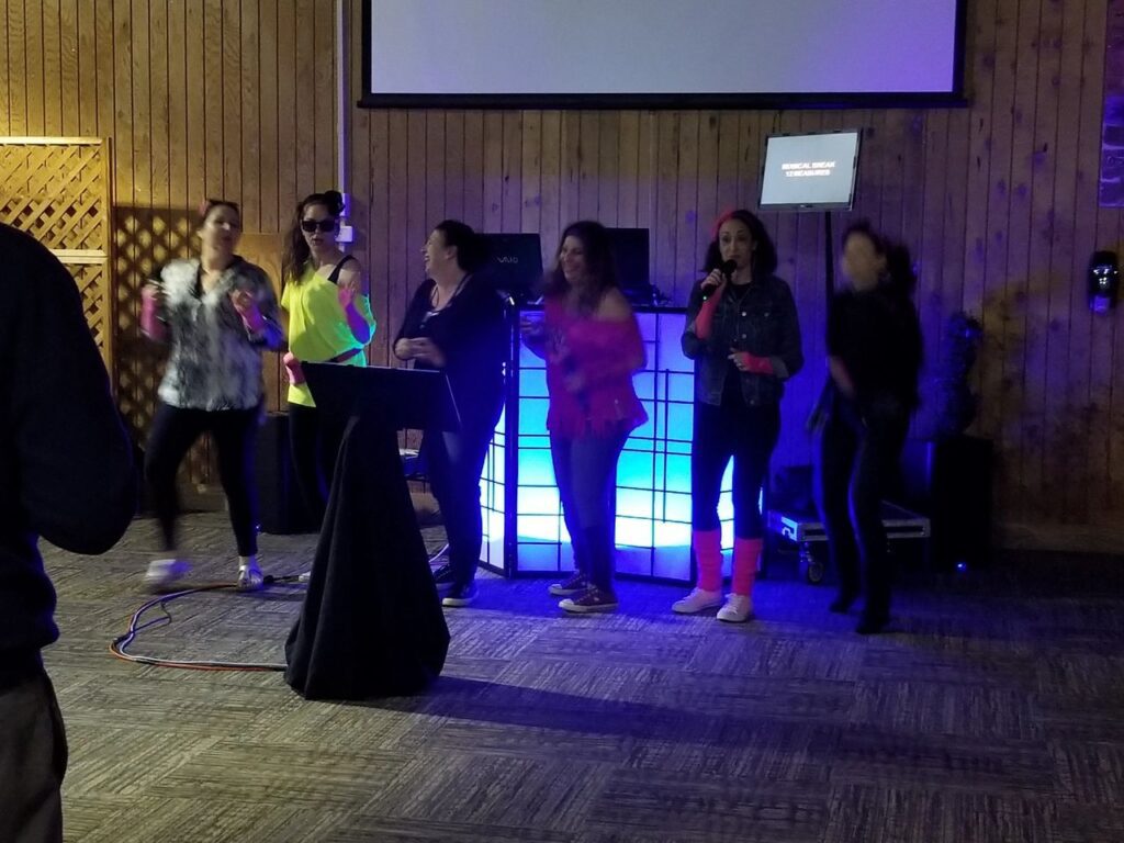 Moms in their 80s theme attire. singing karaoke, dancing on a stage in front of a lighted DJ booth and a karaoke screen in a wood-paneled room.