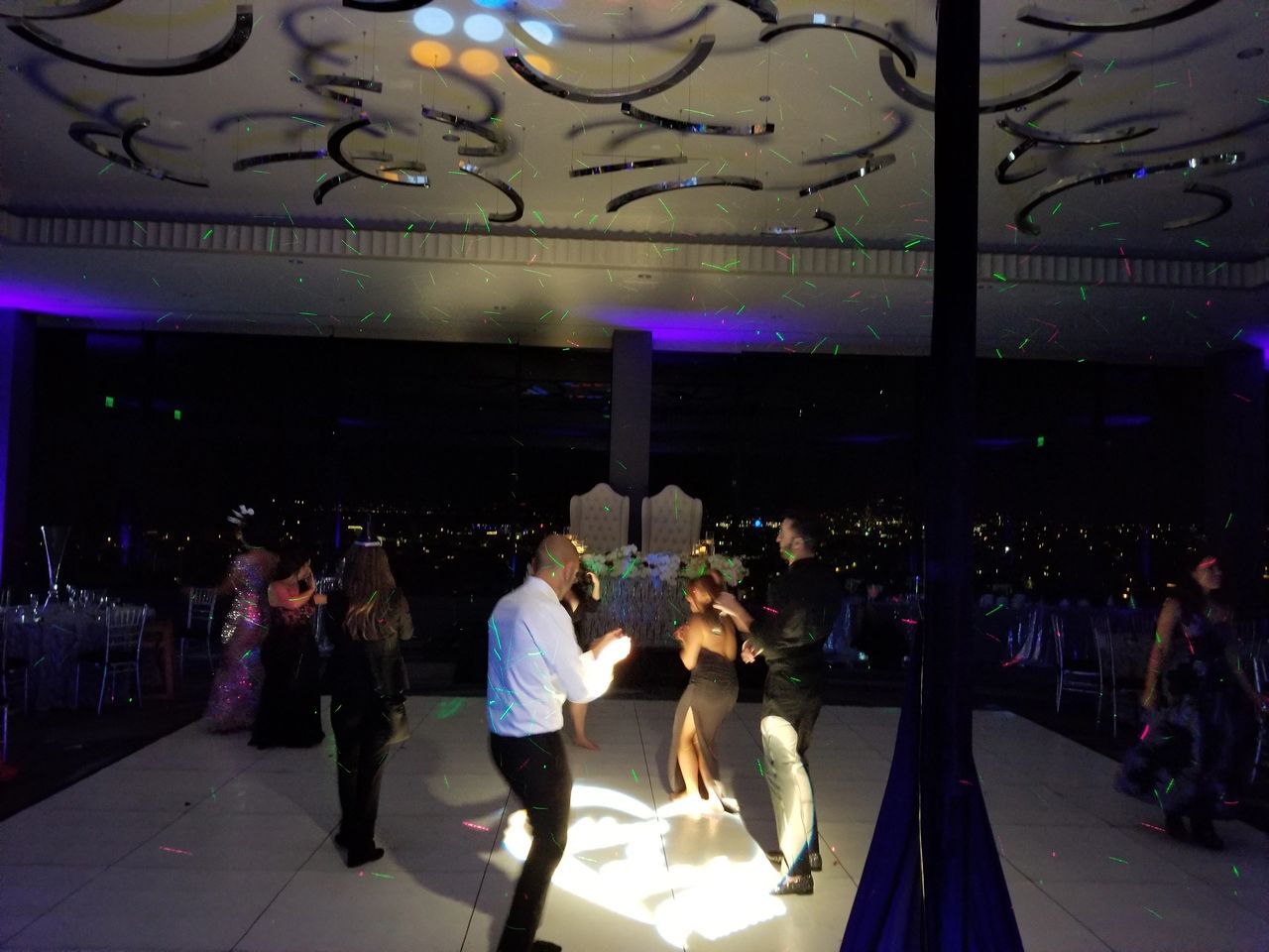 People are dancing on a lit dance floor in a dark room with colorful lights and reflections on the ceiling. Tables and chairs are visible in the background.