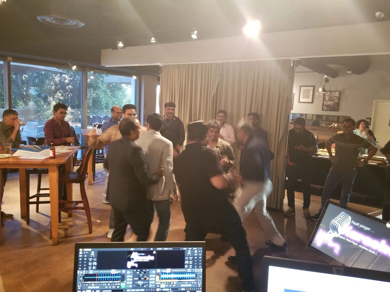 A group of people, some standing and some sitting, gather inside a restaurant; several appear to be involved in an altercation while others watch. Computer monitors are visible in the foreground.