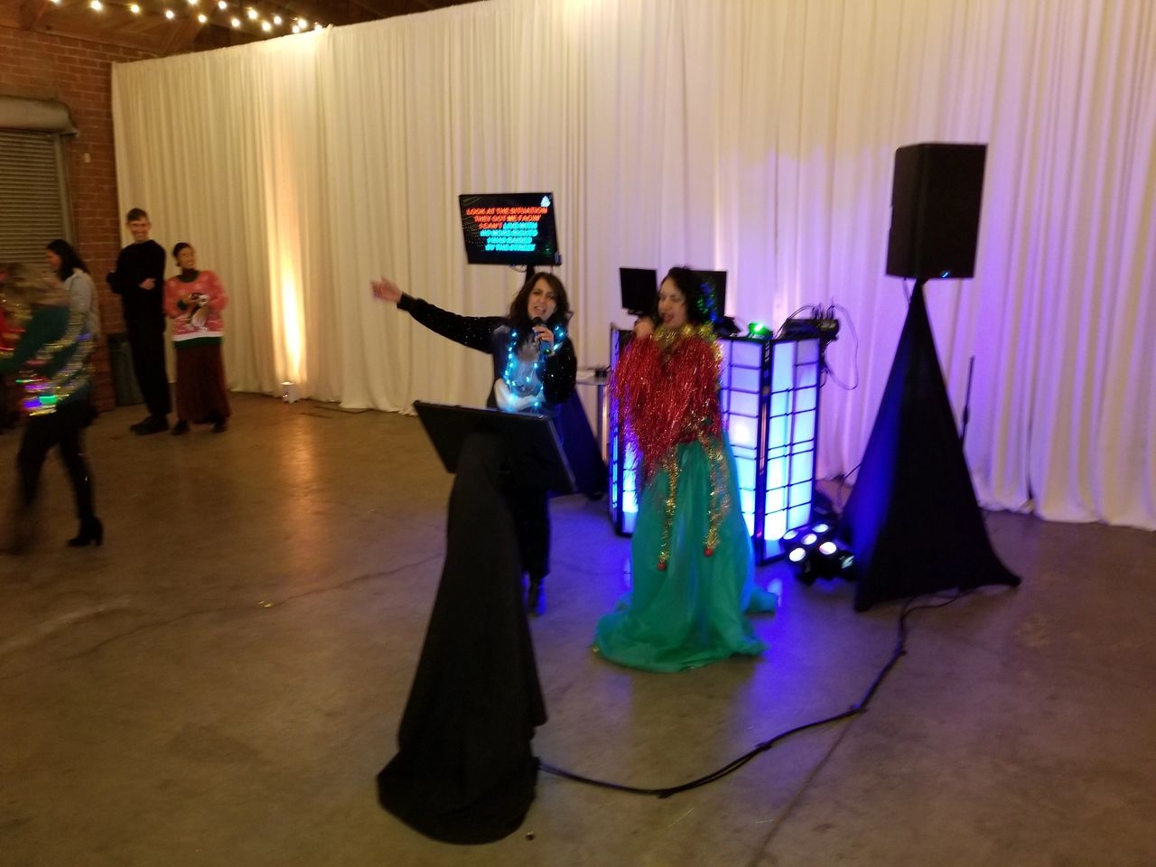 Two people sing karaoke on microphones in a decorated indoor space, with a karaoke screen, speakers, and colored lights behind them. Other people stand and watch.