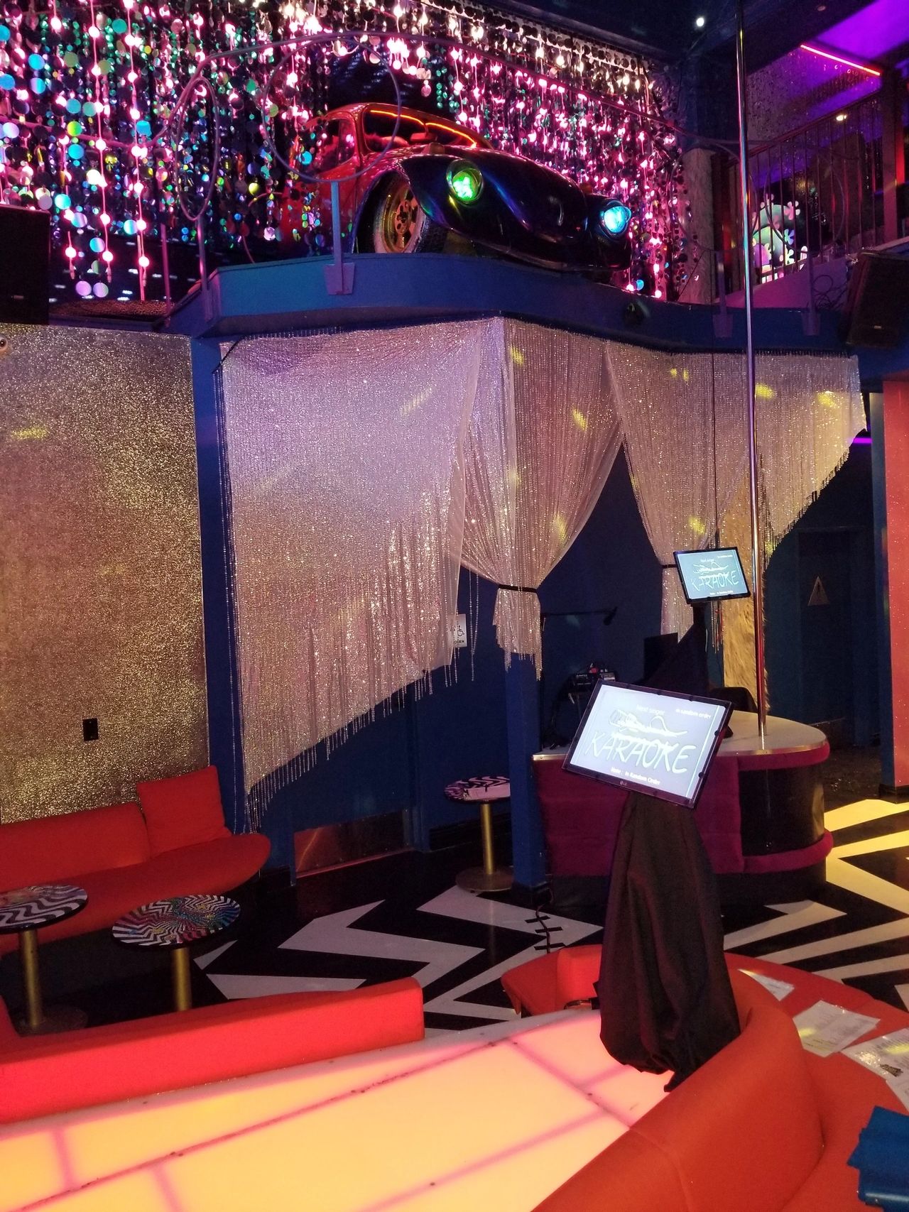 A colorful karaoke lounge with red sofas, a glowing dance floor, a stage area, and a car displayed on an upper ledge amid hanging decorations and shimmering curtains.