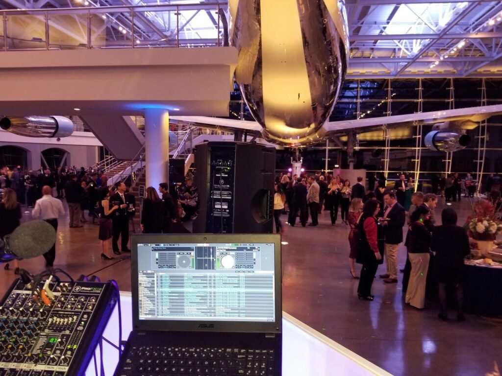 A DJ setup with a laptop and mixer overlooks a large indoor event with people mingling beneath a suspended airplane in a spacious, modern venue.