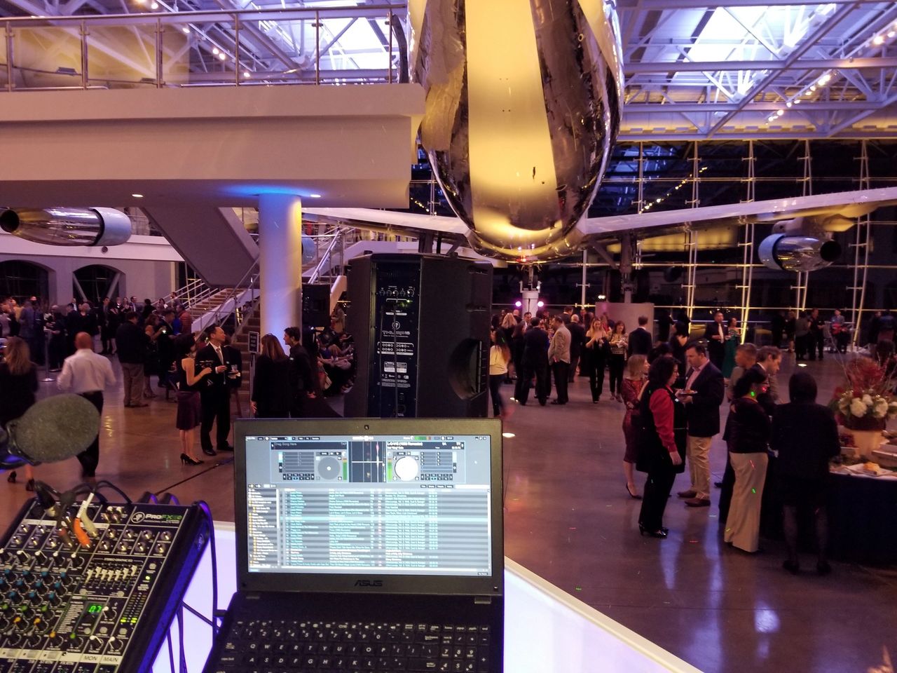 A DJ setup with a laptop and mixer overlooks a large indoor event with people mingling beneath a suspended airplane in a spacious, modern venue.