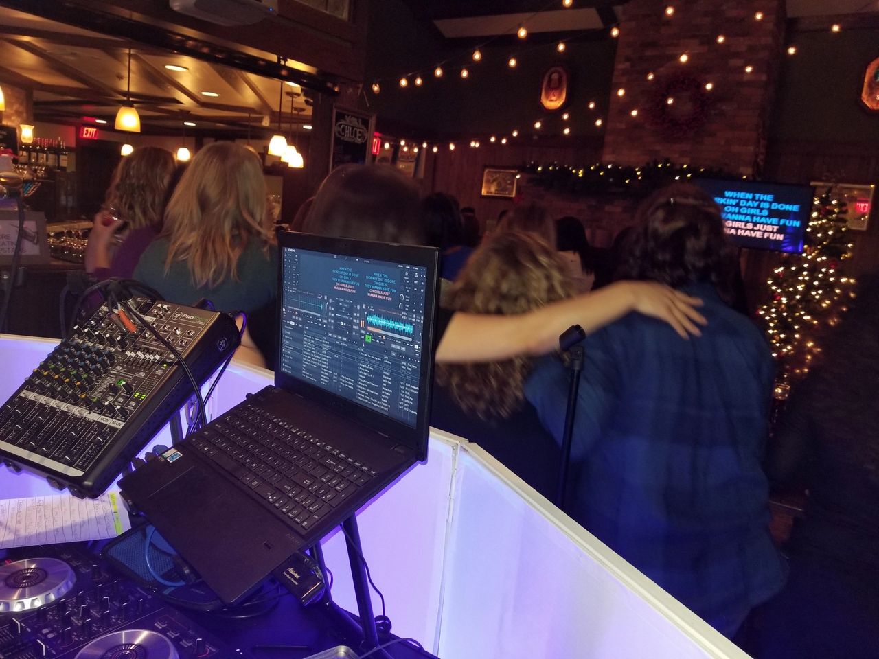 A group of people stand with arms around each other at a karaoke night in a dimly lit bar; a DJ setup and laptop are visible in the foreground.
