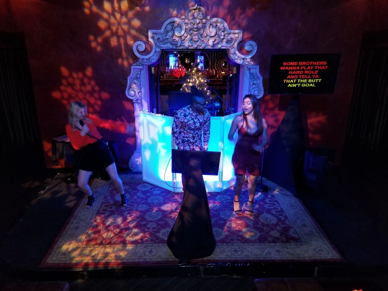 Three people perform karaoke on a stage with ornate decor and colorful lighting; lyrics are displayed on a screen to the right.
