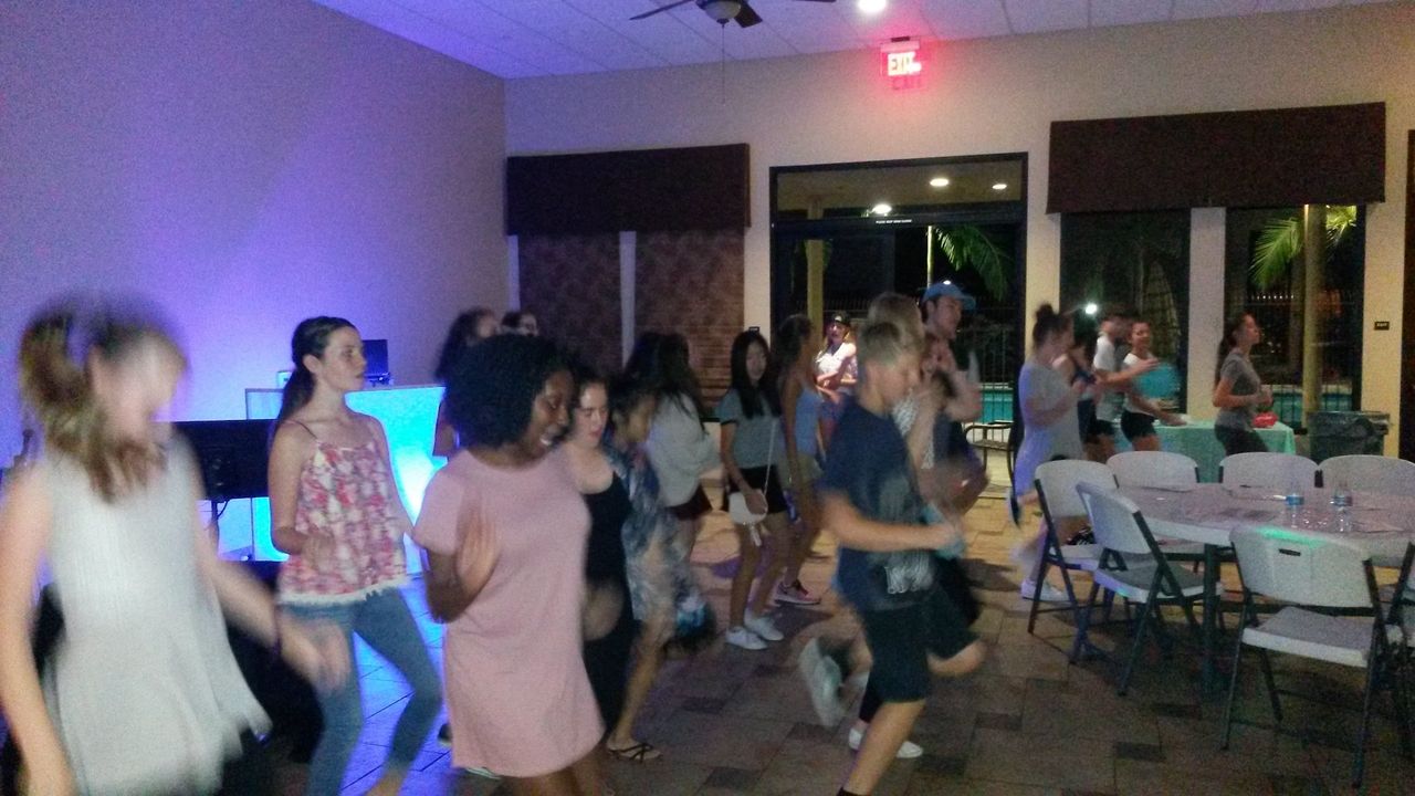 A group of people dance together in a room with tables and chairs; some are blurred due to movement. The room is lit with blue and white lighting.