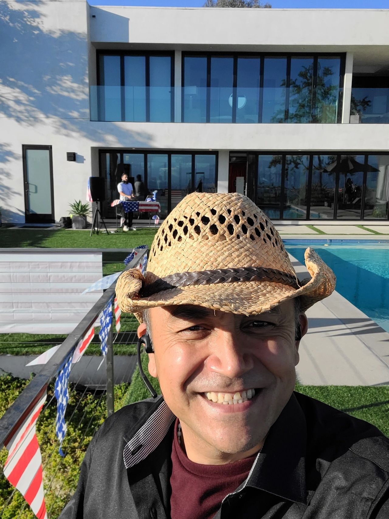 A man wearing a straw hat smiles for a selfie in front of a modern house with large windows, a pool, and outdoor decorations.
