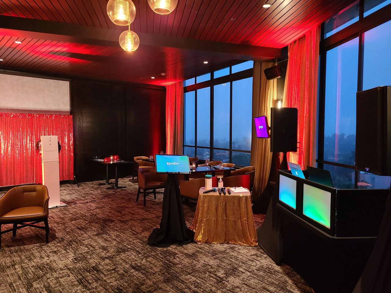 A modern lounge with floor-to-ceiling windows, red ambient lighting, small tables, chairs, a DJ booth, and digital screens set up for an event.