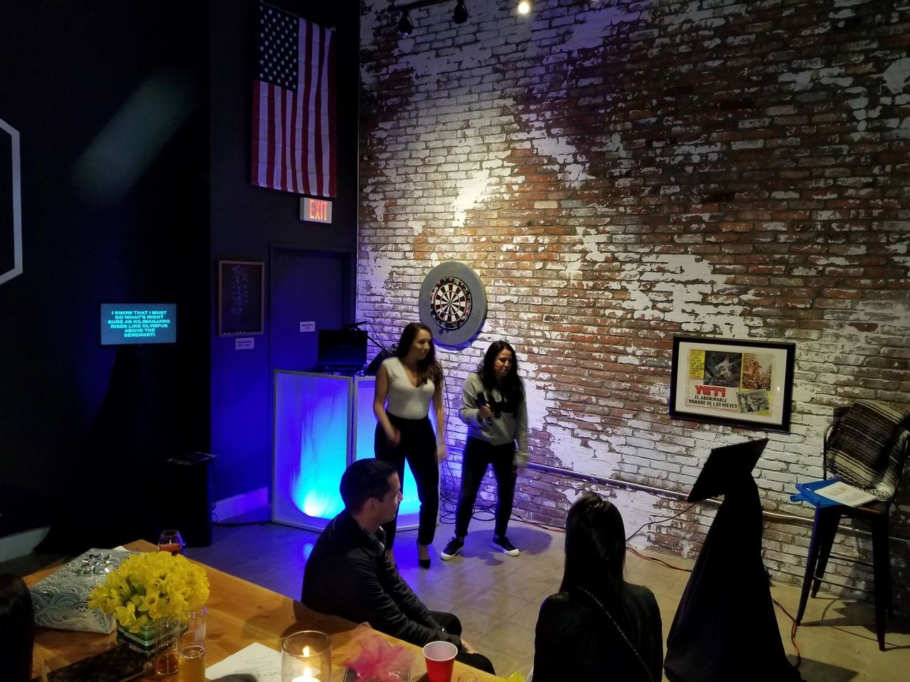Two women perform karaoke in a dimly lit room with a brick wall, dartboard, and American flag; several people are seated and watching.