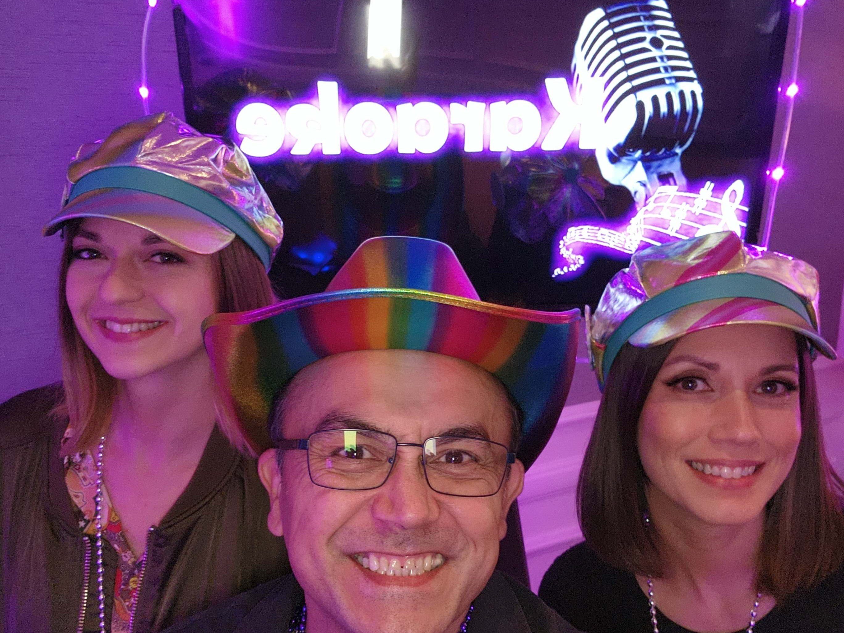 Karaoke Dj Services team members. Three people wearing colorful hats smile in front of a neon "Karaoke" sign and microphone decoration, celebrating in a festive setting