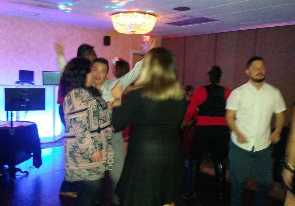 A group of people are dancing and socializing in a dimly lit room with a chandelier and DJ setup.