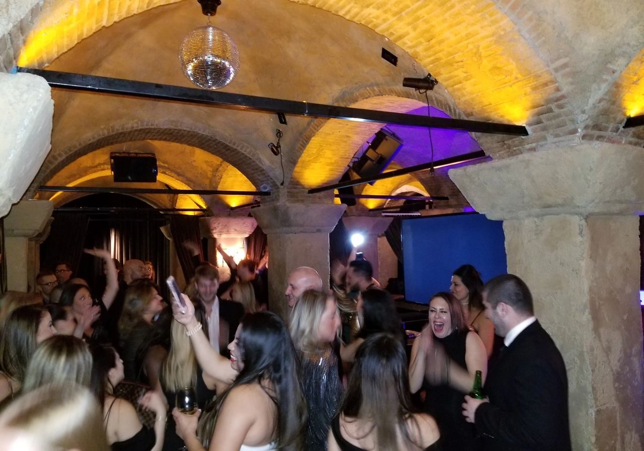 Entrepreneur's Karaoke Dj Party. Guests dressed in formal attire are dancing and socializing at a lively party in a stone-arched room with yellow lighting and a disco ball.