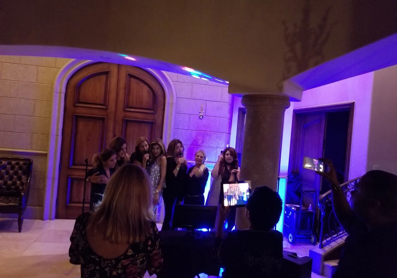 A group of women sing together at a karaoke setup in a room with stone walls and wooden doors, while others watch and record with their phones.