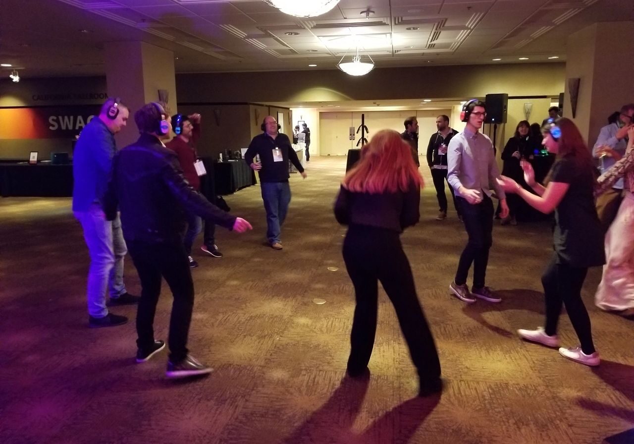 A group of people wearing headphones dance in a dimly lit room with brown carpet and overhead lights, participating in a silent disco event.