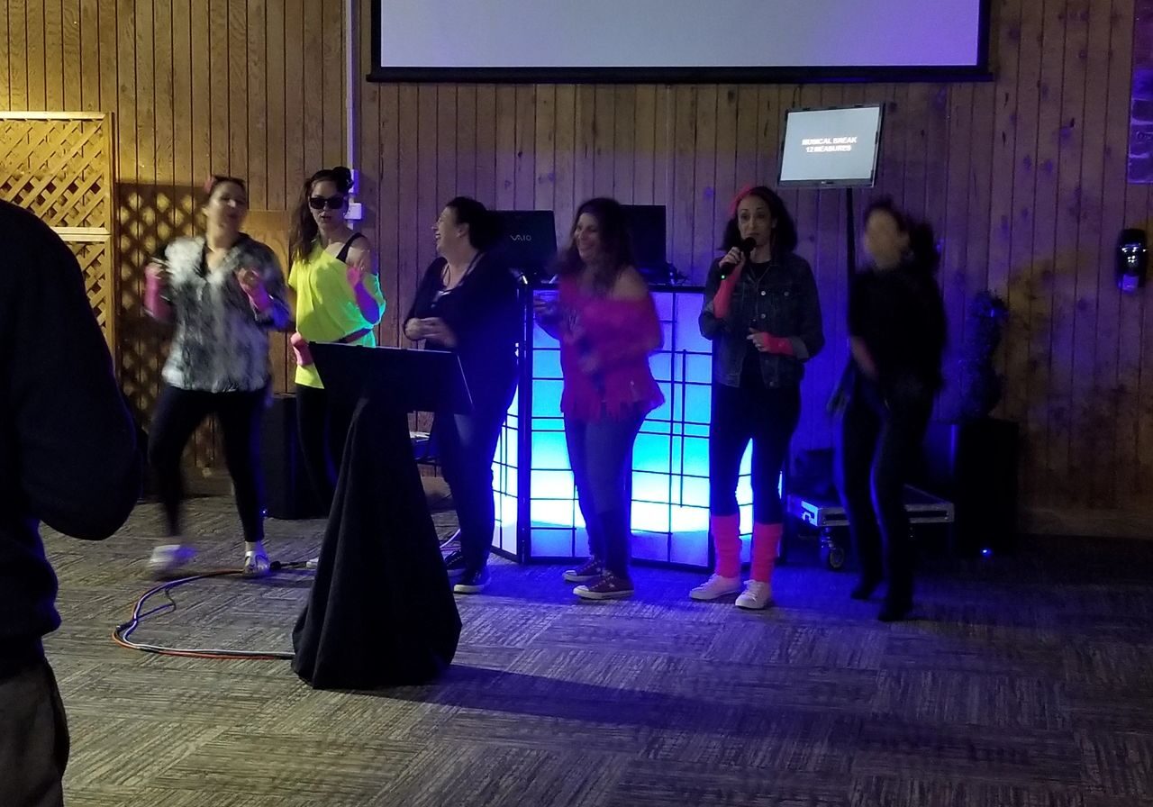 Moms in their 80s theme attire. singing karaoke, dancing on a stage in front of a lighted Party DJ booth and a karaoke screen in a wood-paneled room.