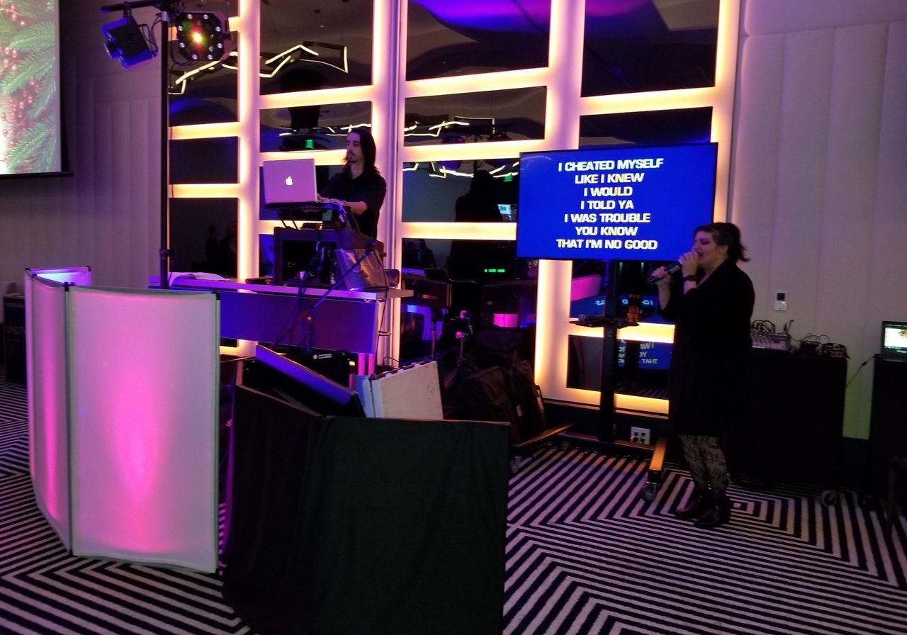 A woman sings karaoke while another operates a laptop at a DJ booth; song lyrics are displayed on a screen beside her in a room with colorful lighting.