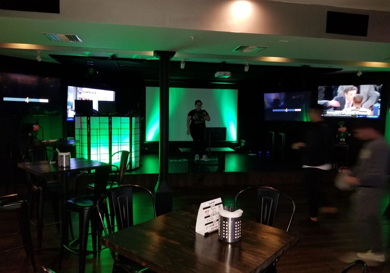 Dimly lit bar with empty tables, a central stage with green lighting, a person on stage, and multiple TV screens on the walls.