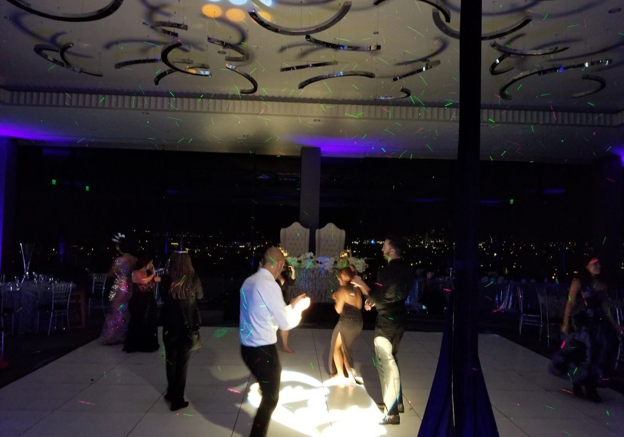 People are dancing on a lit dance floor in a dark room with colorful lights and reflections on the ceiling. Tables and chairs are visible in the background.