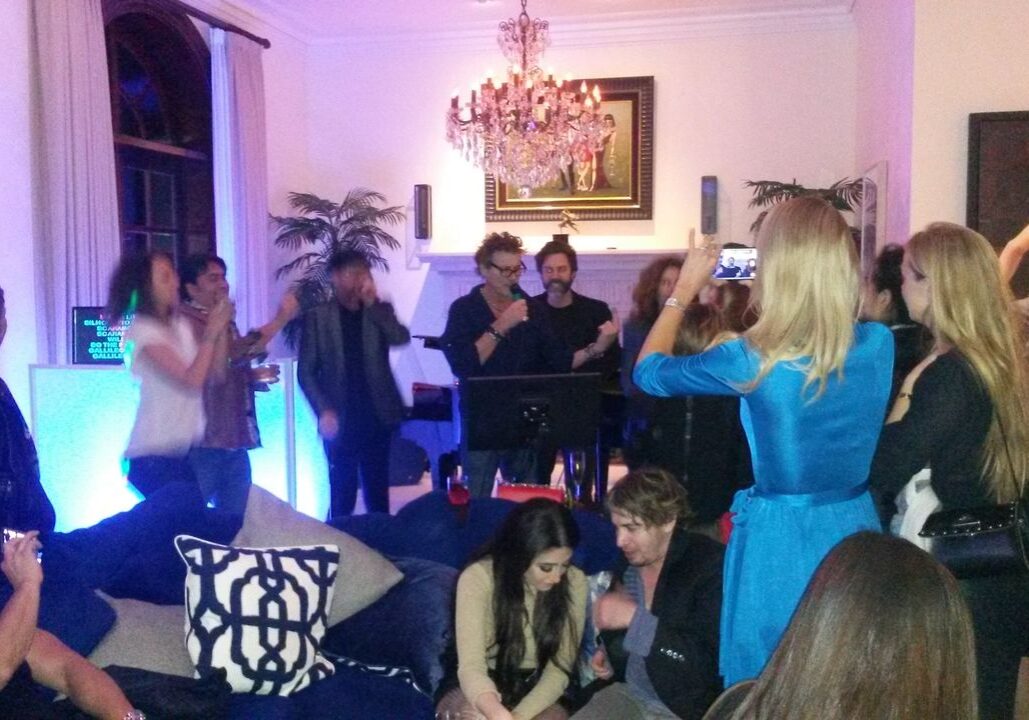 A group of people gather in a living room at night, some singing karaoke under a chandelier, while others watch, sit, or take photos.