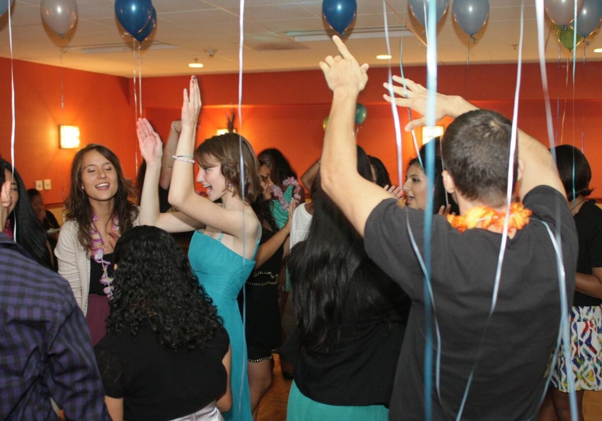 People are dancing and celebrating at an indoor party decorated with balloons and streamers.