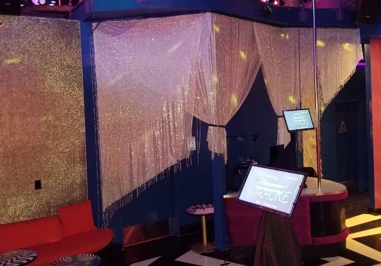 A colorful karaoke lounge with red sofas, a glowing dance floor, a stage area, and a car displayed on an upper ledge amid hanging decorations and shimmering curtains.