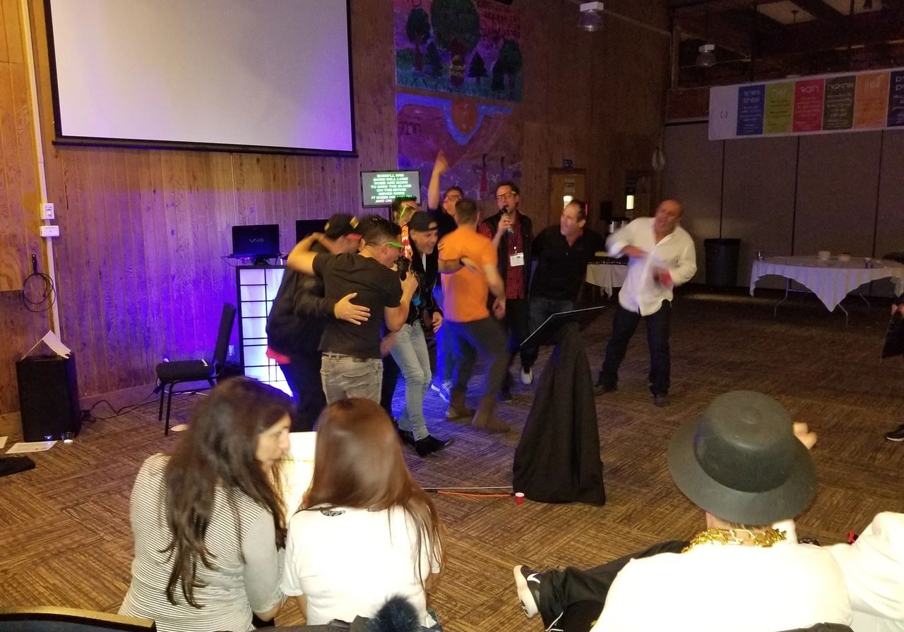 80s theme party. projector screen, some hugging and celebrating, while others sit and watch in a warmly lit room with wood-paneled walls.