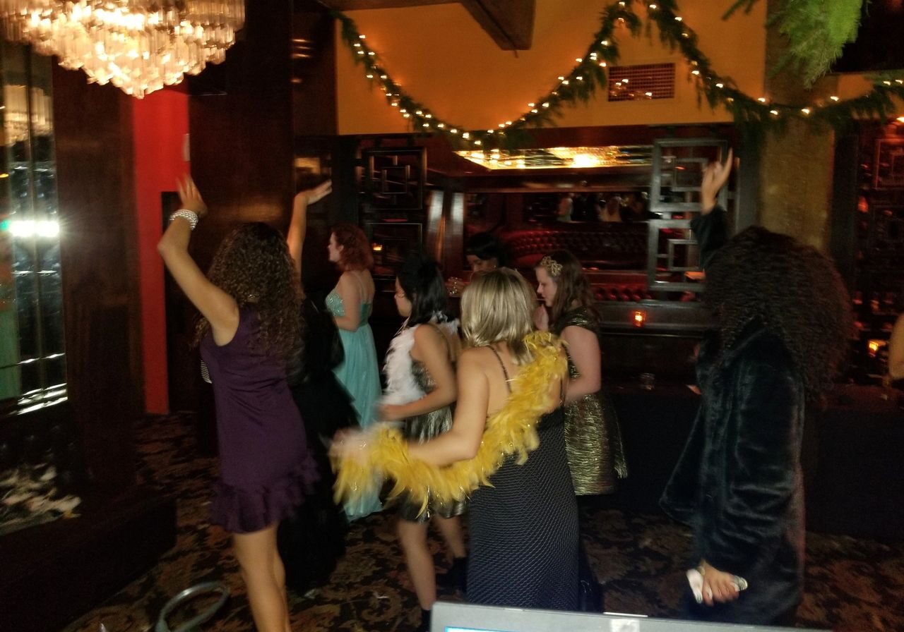 A group of people dressed in formal and festive attire dance and raise their arms in a dimly lit, decorated room with chandeliers and garlands.