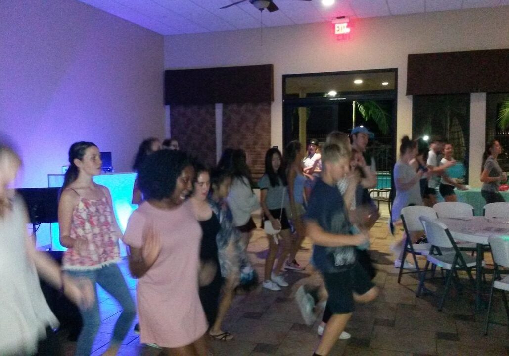 A group of people dance together in a room with tables and chairs; some are blurred due to movement. The room is lit with blue and white lighting.