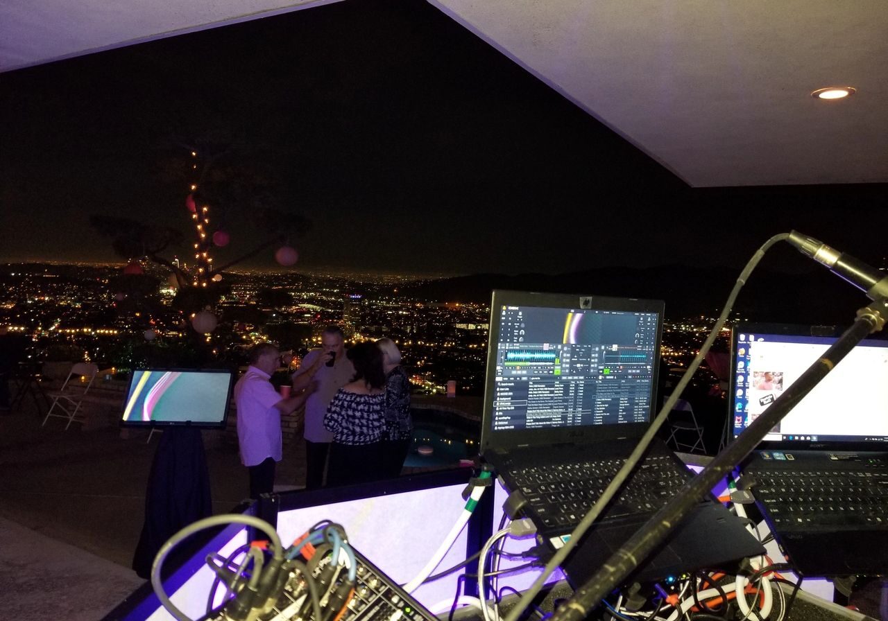 View from a DJ booth overlooking a city at night, with a laptop, cables, and microphones in the foreground and people dancing in the background.