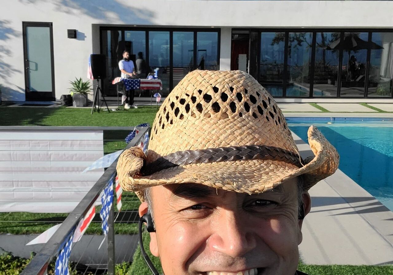 A man wearing a straw hat smiles for a selfie in front of a modern house with large windows, a pool, and outdoor decorations.