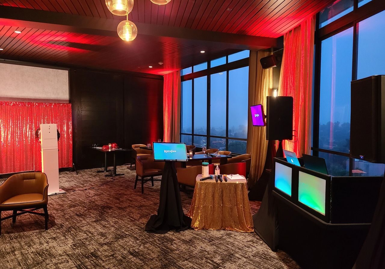 A modern event space with red lighting, tables and chairs, a DJ booth with colorful screens, a photo booth, and floor-to-ceiling windows overlooking a cityscape.